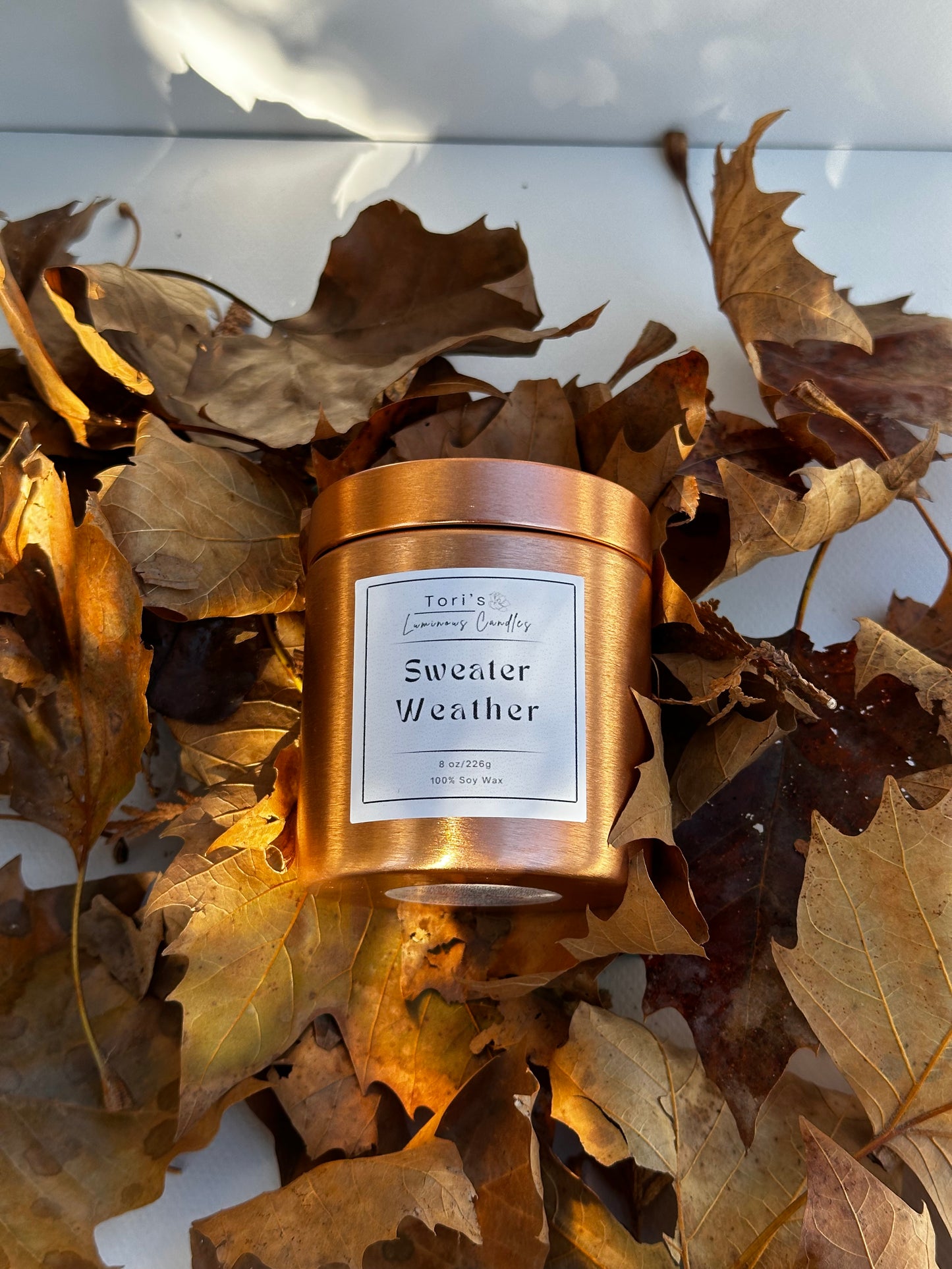 A cozy candle labeled “Autumn Orchard Glow” sits beside fresh apples, cinnamon sticks, and pine sprigs, with cranberries scattered on a rustic wooden table, evoking the warmth and freshness of fall.
