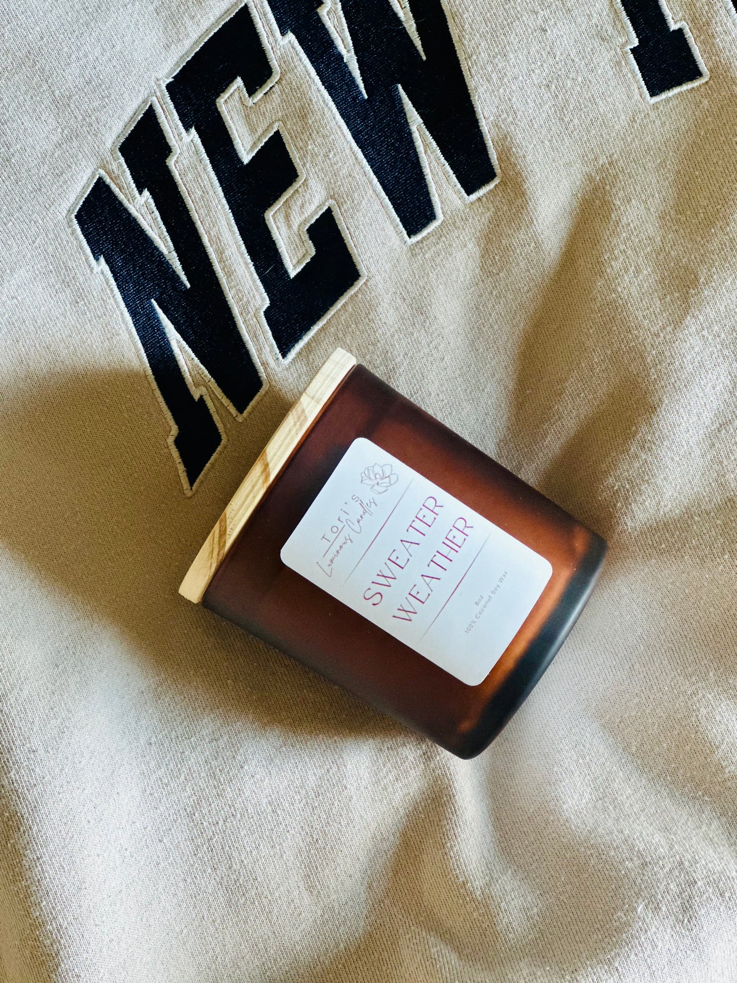 A cozy candle labeled “Autumn Orchard Glow” sits beside fresh apples, cinnamon sticks, and pine sprigs, with cranberries scattered on a rustic wooden table, evoking the warmth and freshness of fall.