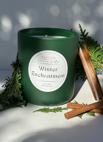 A Winter Enchantment soy candle surrounded by cozy winter accents. The label highlights notes of citrus, pine, pear, juniper, and sage. The scene evokes a snowy forest feeling with a serene, winter wonderland aesthetic.