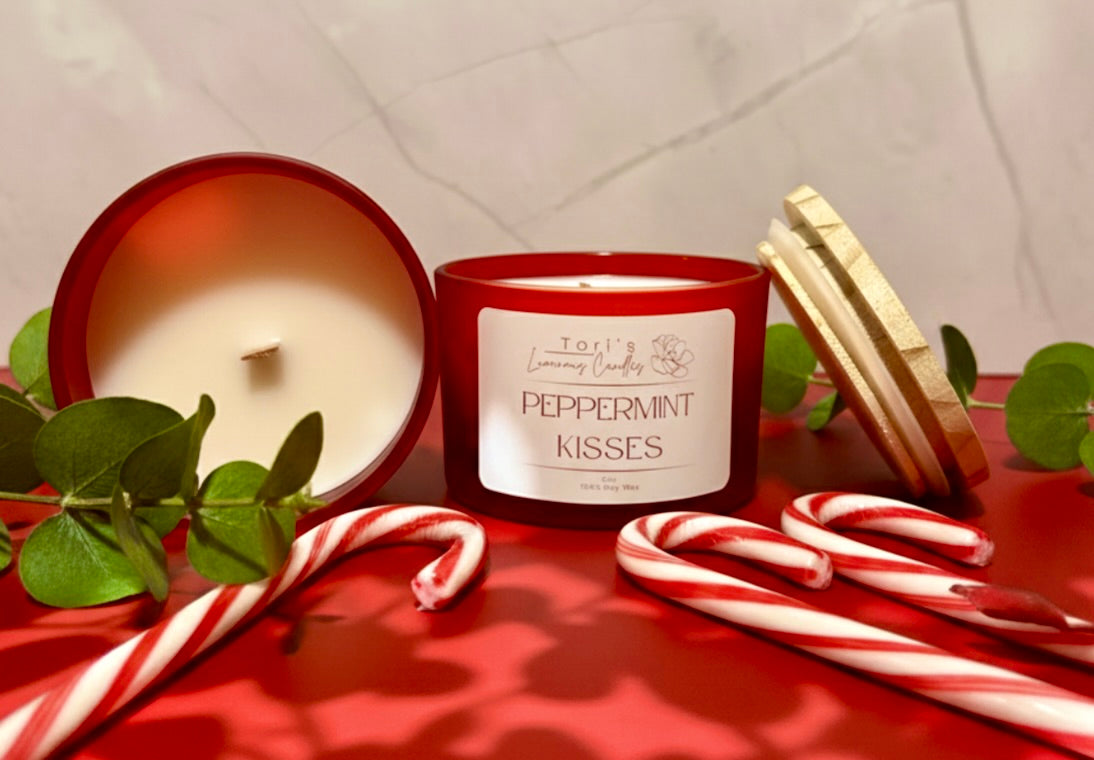 Peppermint Kisses candle lit on a clean winter-themed surface, surrounded by red-and-white peppermint candies and soft eucalyptus sprigs. The flame glows brightly, creating a fresh, uplifting holiday feel reminiscent of candy canes and winter air.