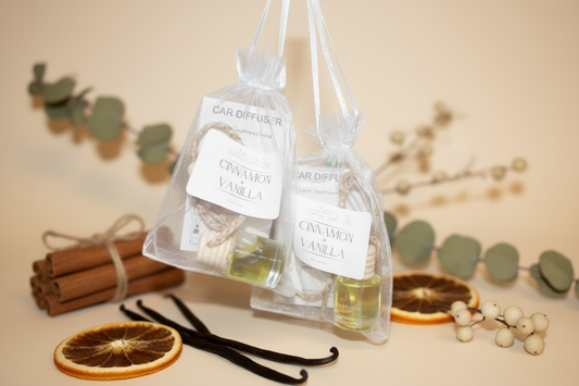 A close-up of a small glass car diffuser with a wooden lid, filled with clear fragrance oil. The label reads 'Cinnamon & Vanilla.' It is hanging from a rearview mirror with a soft-focus view of a steering wheel and road in the background.